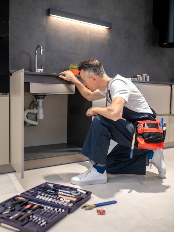 Household repair. Middle-aged man in blue overalls knelt down with hand touching sink carefully examining pipes in kitchen at home